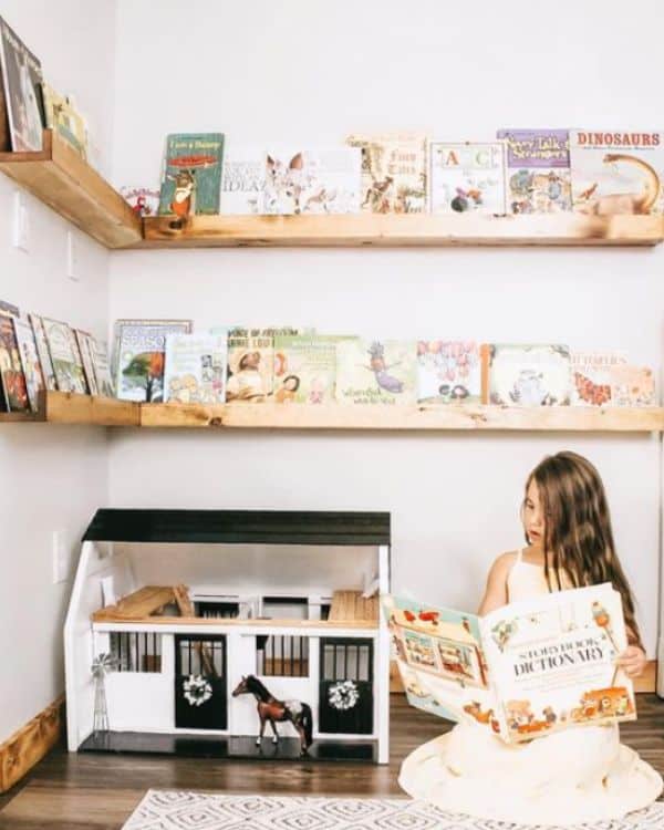 Image of a book corner in a child's room. The wooden ledge shelves wrap around the corner of the room holding books. A child sits on the floor reading next to her toy farmhouse. 