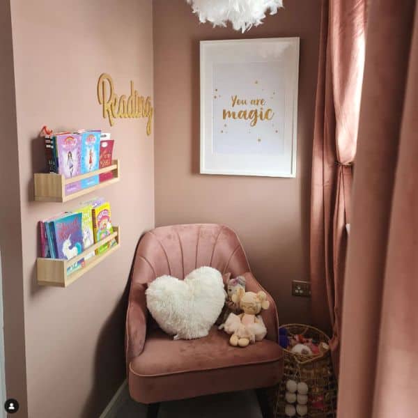 A cozy kids book nook in the corner of a room. The walls are a beautiful blush pink, the chair is pink, the curtains are pink, and a couple of wooden floating shelves are on the wall holding books to read. 