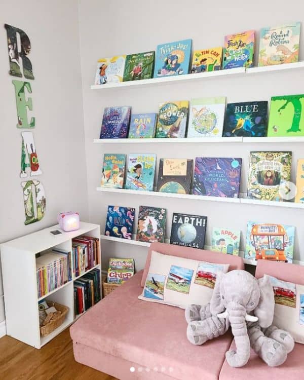 Cute Reading Corner For Kids - A Pink Nugget Play Couch sits in front of a wall with white ledge shelves holding various childrens books. A cube shelf is on the adjacent wall with a "READ" sign hanging above it. 