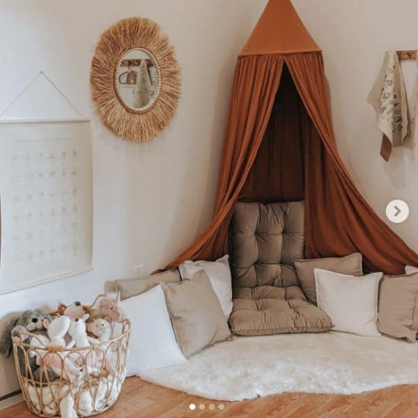 Kids Reading Nook Ideas - This image has a white fluffy rug in the corner with various white and tan throw pillow. A burnt orange canopy hangs from the ceiling overhead. 