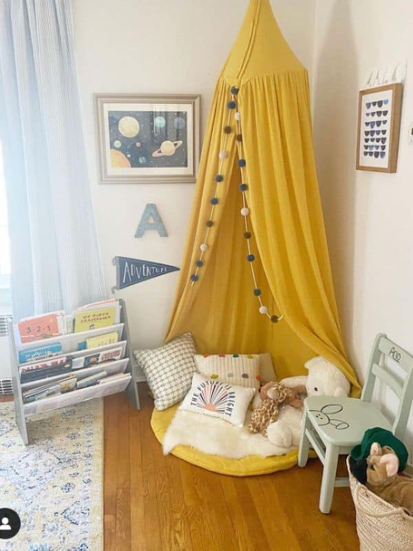 Toddler Reading nook idea - A yellow canopy hangs from the ceiling over a yellow round cushion on the ground with a variety of throw pillows on top of it. Next to the cushions is a tiered shelf holding children's books to read. 
