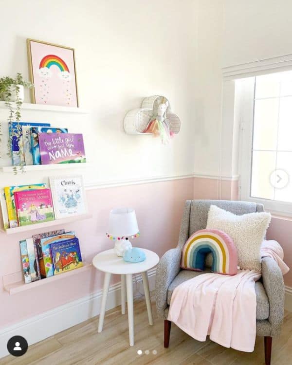 Nursery Reading Nook Idea - a comfy gray chair in the corner of the room with a pink blanket draped over it. Next to the chair is a small white side table and floating ledge shelves holding books. 