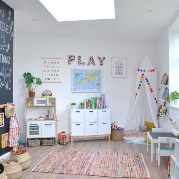 An image of a child's playroom. In the corner of the playroom is a cozy reading area with various pillows and a sheer canopy with rainbow pom poms hanging from the ceiling. 