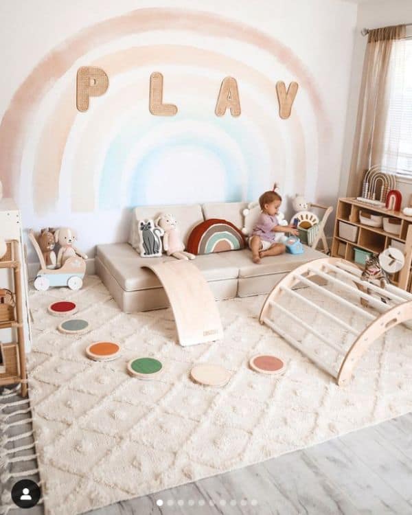 Montessori-inspired playroom - A baby plays in a cozy playroom. A rainbow is painted on the back wall with the Word "Play" spelled out in wooden letters. The playroom includes wooden stepping stones, a tan Nugget play couch, a Pikler arch, and a variety of Montessori toys on a short wooden shelf. 