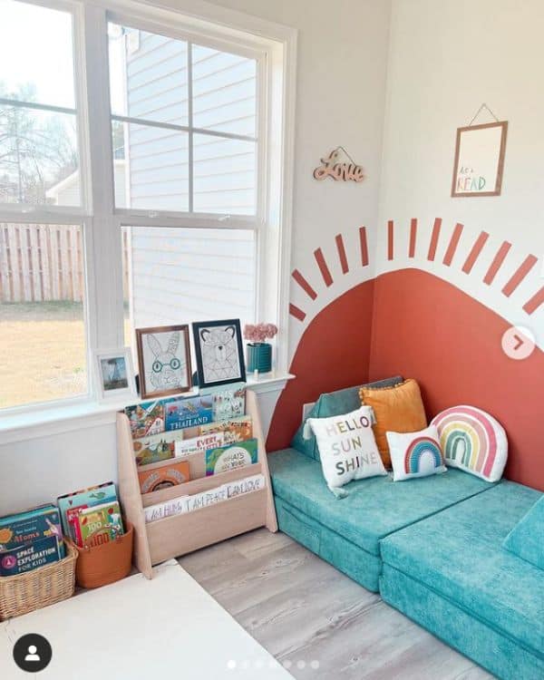 Kids Reading Nook Ideas - A sun mural painted on the wall in the corner. In front of the mural sits a blue Nugget Play couch and a tiered wooden book shelf with children's books to read. 