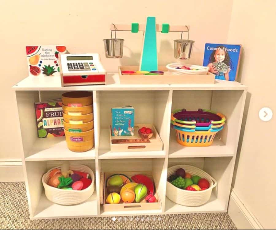 Montessori Playroom ideas - A white 6-cube cube storage shelf sits against a white wall in the corner of the room. On the shelf are a variety of food and grocery toys like shopping baskets, play food, and a cash register. 