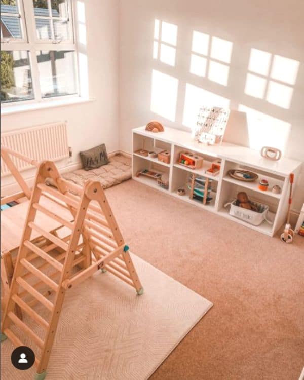 A camera view from above a Montessori playroom shows a short white shelf with wooden toys on it and a wooden pickler triangle climbing structure in the center of the room. 