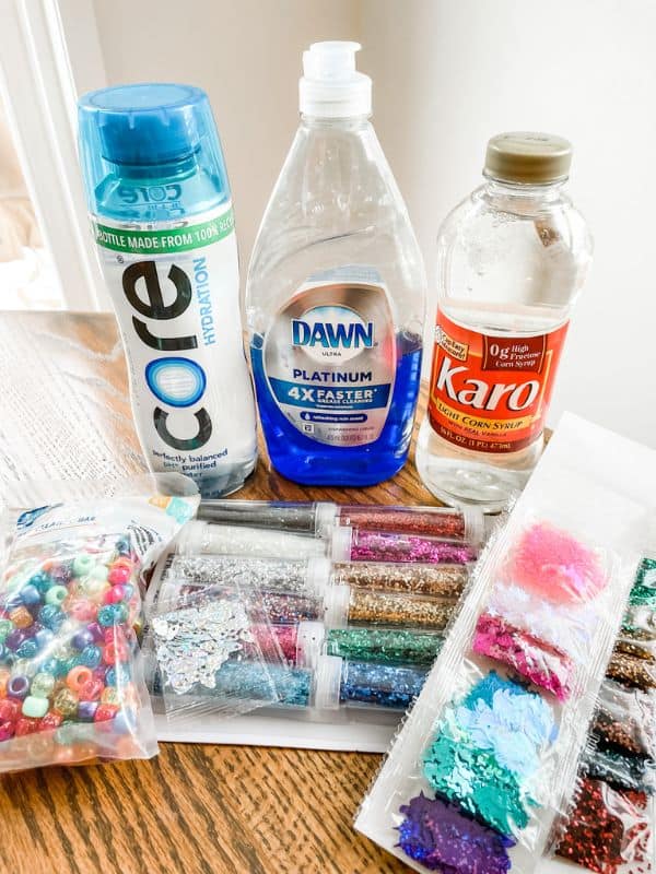 Image of materials used to make diy sensory bottles. The materials include a plastic water bottle, dish soap, clear corn syrup, glitter, and beads. 