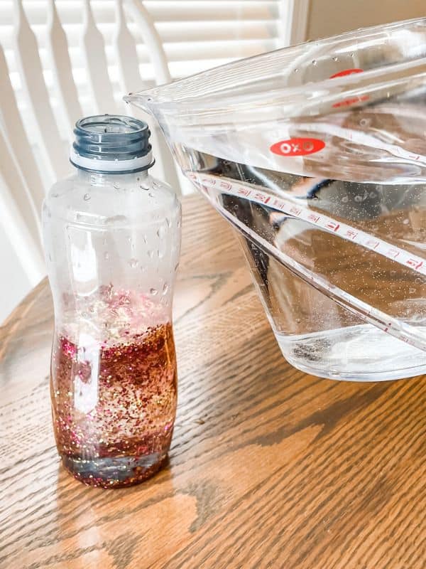 Image of clear plastic water bottle filled a little over halfway with glitter and clear corn syrup and someone pouring warm water into the rest of the bottle to fill it up. 
