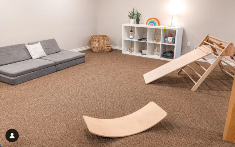 Image of a minimalist playroom with a gray play couch, toy shelf with a few wooden toys, pickler triangle, and a wobble board. 