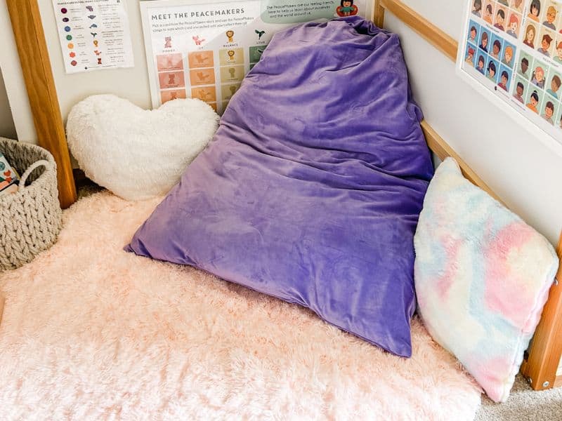 Calming corner with soft pink rug, purple bean bag, rainbow throw pillow, and a heart shaped throw pillow. 
