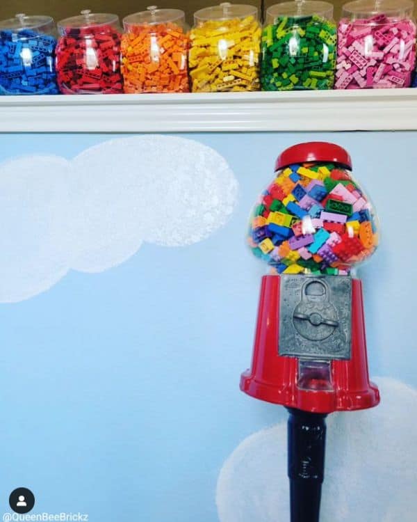 A row of clear plastic containers filled with different colors of Lego pieces, placed on a shelf, with a gumball machine filled with Legos.