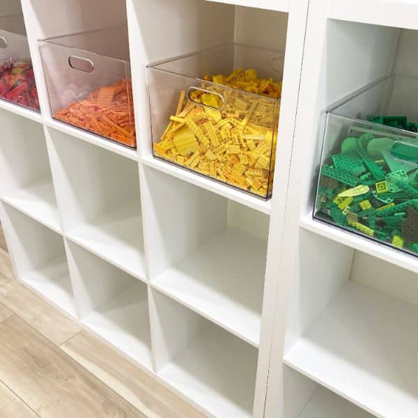 A white cubby shelf with large clear bins filled with Lego pieces sorted by color.