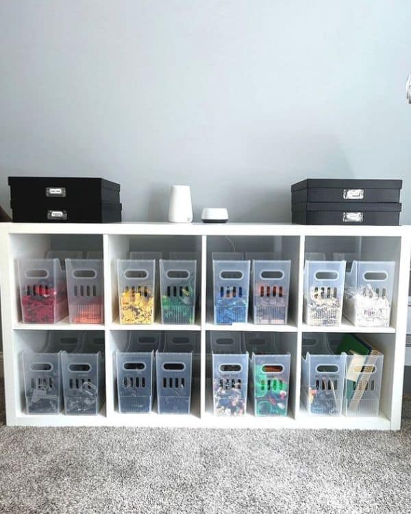 A white cubby shelf unit with clear bins labeled and filled with various colors of Lego pieces.